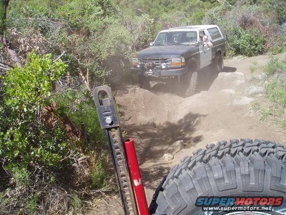 Off-roading in a dark SUV with a white top, kicking up dust on a rocky trail. Another vehicle is visible ahead