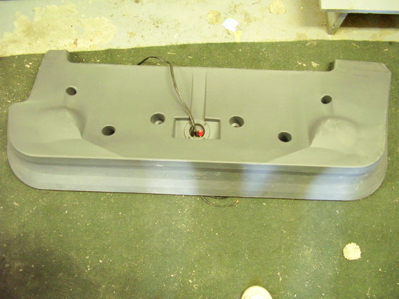 Gray plastic bumper part with circular indentations and a central red light, lying on a green floor