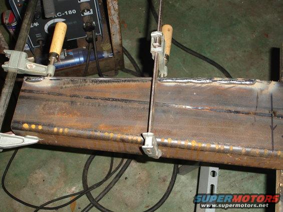 Metal beam being welded, held by clamps, with a welding machine in the background