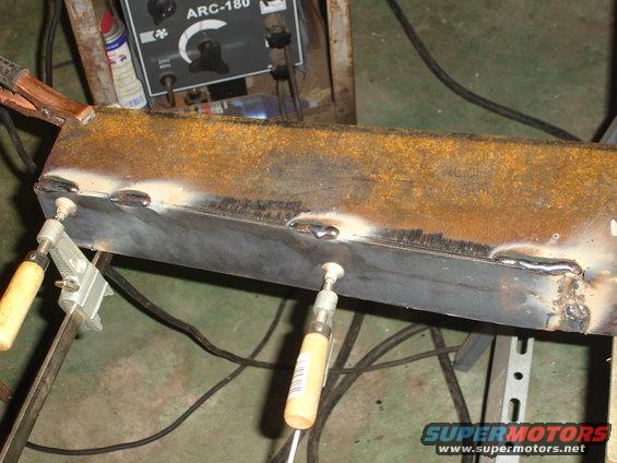 Rusty metal beam being welded, held by clamps, with a welding machine in background