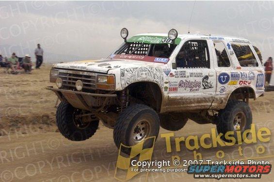 White off-road truck with racing decals, lifted suspension, and large tires, mid-air over a dirt track