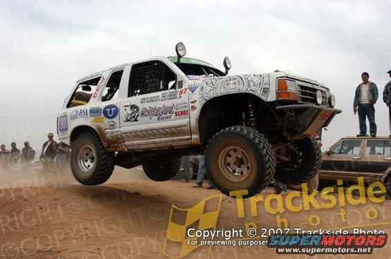 White off-road SUV airborne, modified with large tires and roll cage. Graphics on hood and doors. Another vehicle and spectators visible