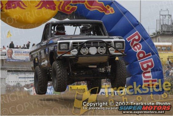 Pickup truck with roll cage and off-road tires mid-air jump, Red Bull arch in background