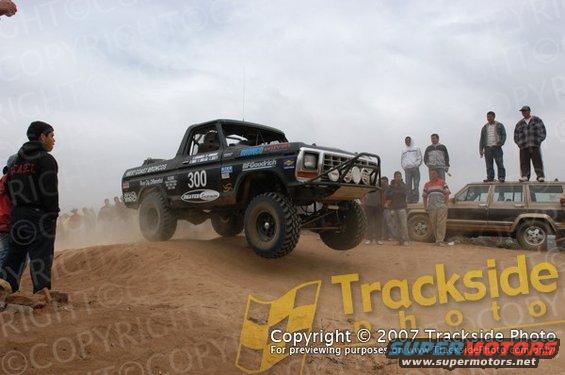 Off-road race truck with number 300 on door, mid-air over dirt mound. Another SUV is parked nearby with people on top