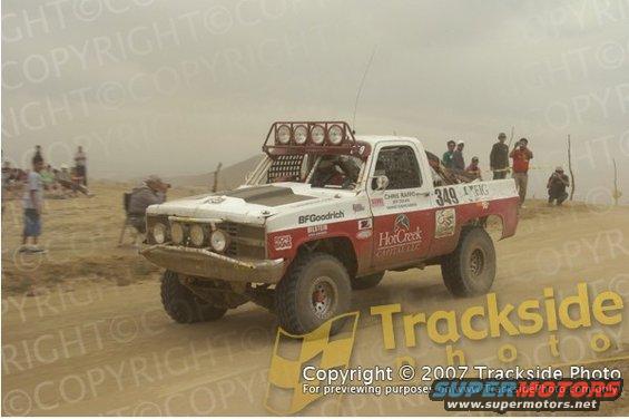 White and red off-road racing truck with large tires and light bar on roof, kicking up dust