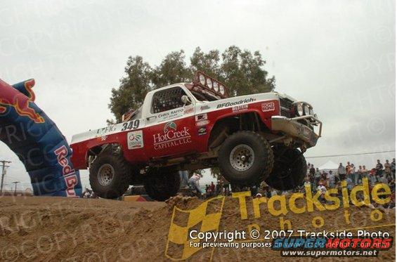 Off-road racing truck with number 349 airborne over a dirt jump, Red Bull arch visible on left