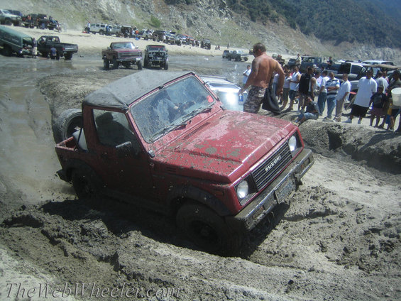 Red Suzuki Samurai stuck in mud, front right tire submerged. Grille reads SUZUKI. Body style suggests mid-1980s