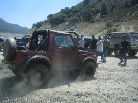 Red Suzuki Samurai kicking up dust off-road with other vehicles and people nearby
