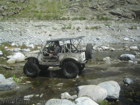 Mud-splattered white Suzuki Samurai driving through a rocky stream bed