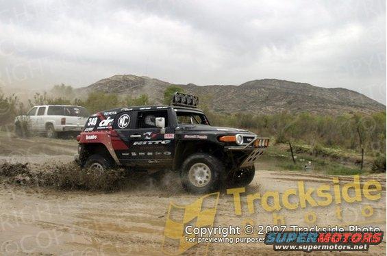 Black Toyota FJ Cruiser driving through mud, with a white SUV in the background