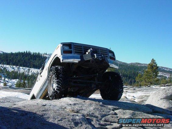 White off-road SUV with large tires climbing over rocks, front grille visible