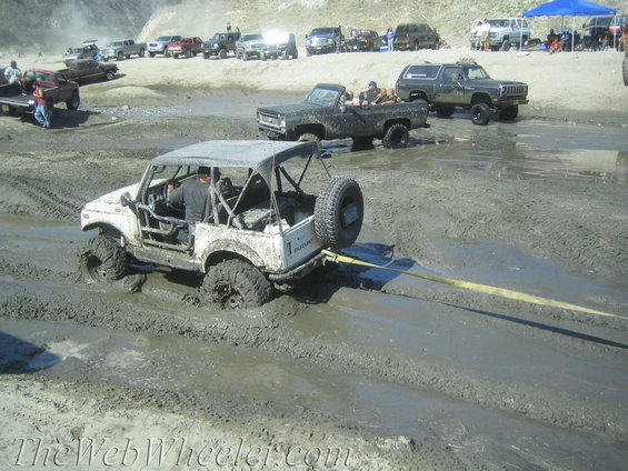 White Suzuki Samurai stuck in mud, being towed by yellow strap. Spare tire mounted on rear