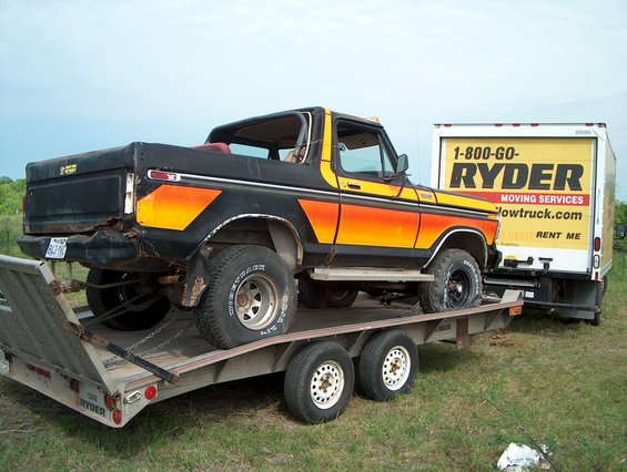 Black 1978 Ford Bronco with orange and yellow stripes on a trailer, towed by a Ryder truck
