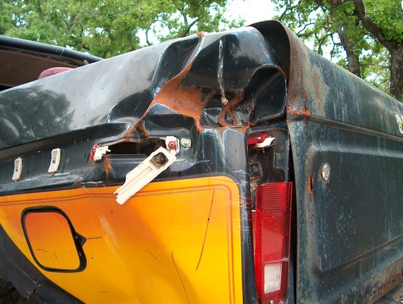 rear corner of a dark-colored truck with severe rear-end damage, showing rust and a broken taillight assembly