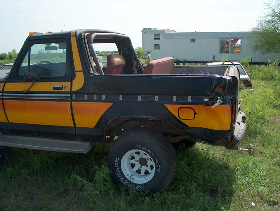 1978 Ford Bronco with black and orange paint, viewed from the rear quarter. The vehicle is in a grassy area with a trailer and trees in the background
