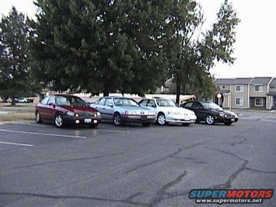 Four sedans parked in a lot with trees and buildings in the background