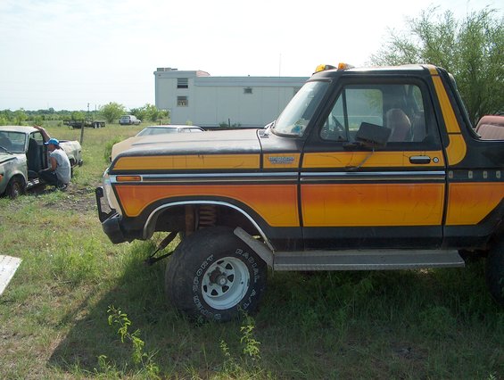 Side view of a 1979 Ford Bronco with orange and yellow stripes, parked in a junkyard