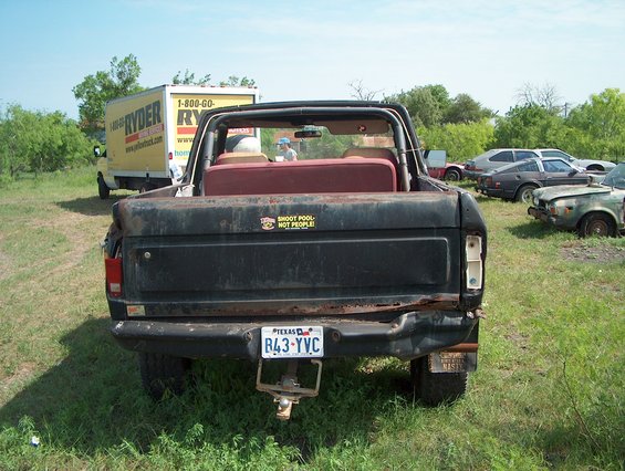 1979 Ford Bronco rear view, showing rust and damage. Other vehicles and a Ryder truck are in the background