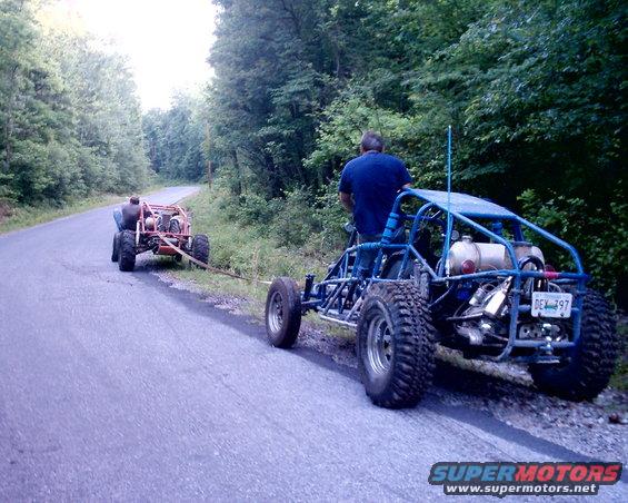 Blue dune buggy towing a red dune buggy on a paved road with trees on the side. License plate reads DEX 797