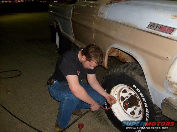 Guy kneeling by a lifted truck, airing up the front passenger tire. The truck has a two-tone paint job and large off-road tires
