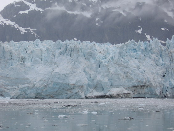 Massive glacier face with jagged ice formations, meeting a body of water with ice floes. Snow-capped mountains rise in the background under a cloudy sky