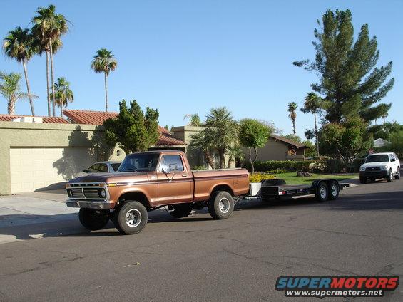1977 Ford F-150 towing an empty car trailer down a residential street. White SUV visible in background