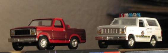 Two toy trucks on a shelf: a red pickup and a white police SUV with lights. The police SUV has text and a badge