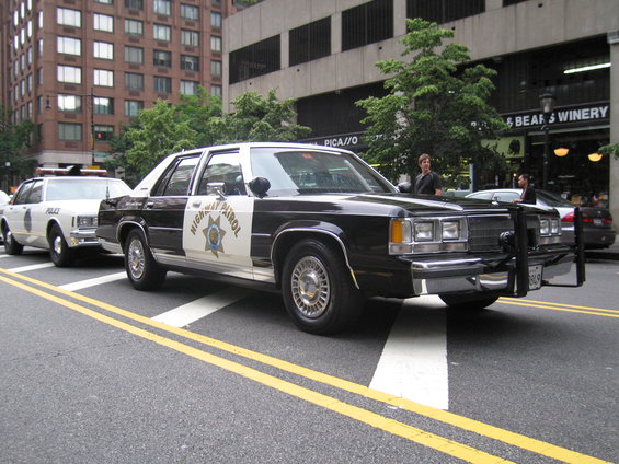 Black and white police car with 'Highway Patrol' on the side, parked on a city street next to another police car
