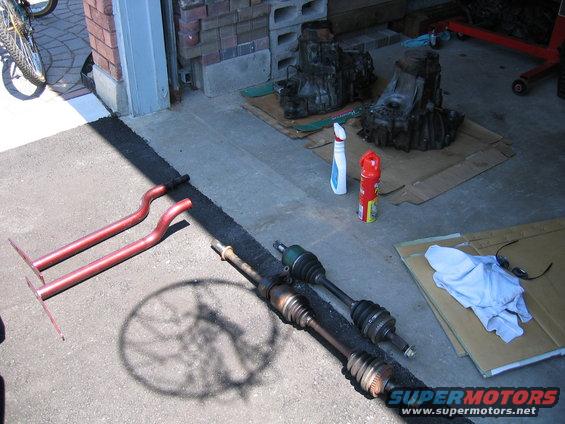 two drive axles and red exhaust pipes on driveway, with two transmissions and spray cans in background