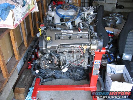 Mazda engine on a red engine stand, with various hoses and wires attached. Some boxes and car seats are visible in the background