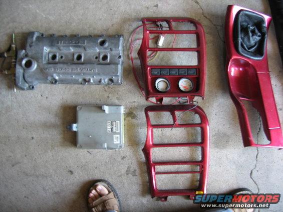 Mazda engine valve cover, ECU, and red interior trim pieces laid out on concrete
