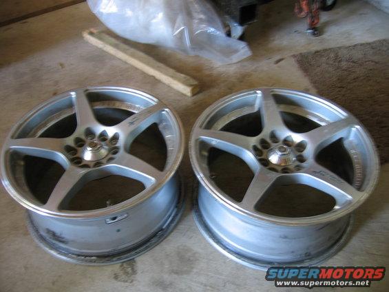 Two silver 5-spoke aftermarket wheels sit on a concrete floor