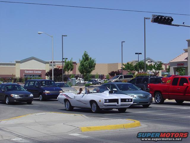 White convertible muscle car with dual headlights and chrome trim driving on a sunny day. Other cars and buildings are visible