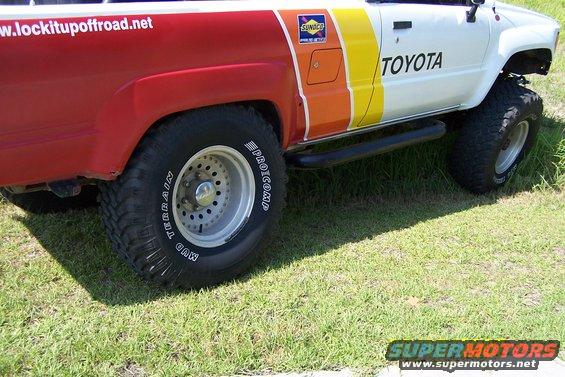 Side view of a Toyota pickup with red rear fenders and a colorful stripe. It has off-road tires and a website address on the fender