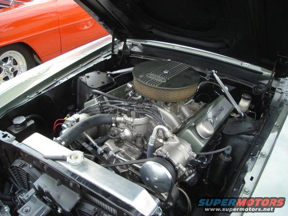 Engine bay of a classic car with a V8 engine, chrome parts, and an orange car visible in the background