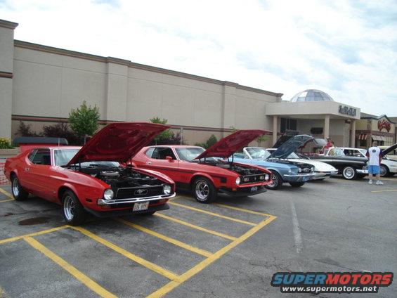 Several classic Ford Mustangs parked in a lot with hoods up. One red Mustang is in the foreground