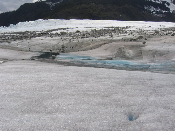 Glacier surface with blue meltwater pools and rocky debris, mountains in background