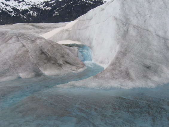 Glacier ice with blue meltwater streams and pools, snow-capped mountains in background