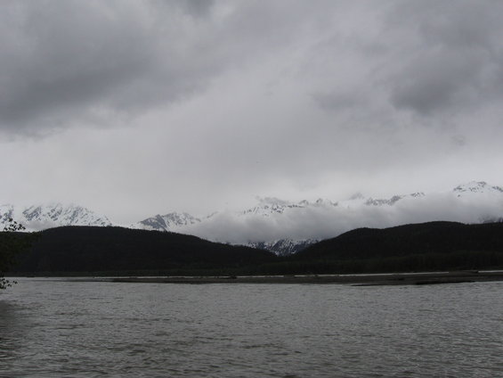 Overcast sky above snow-capped mountains and dark forested hills, with a wide river in the foreground