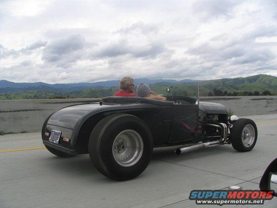 Black hot rod roadster with exposed engine and large rear tires, driving on highway