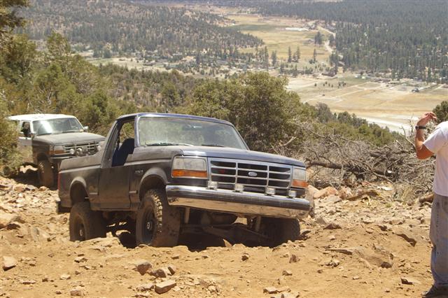Dark blue Ford Bronco and white Ford Bronco driving on a dirt trail with a valley in the background