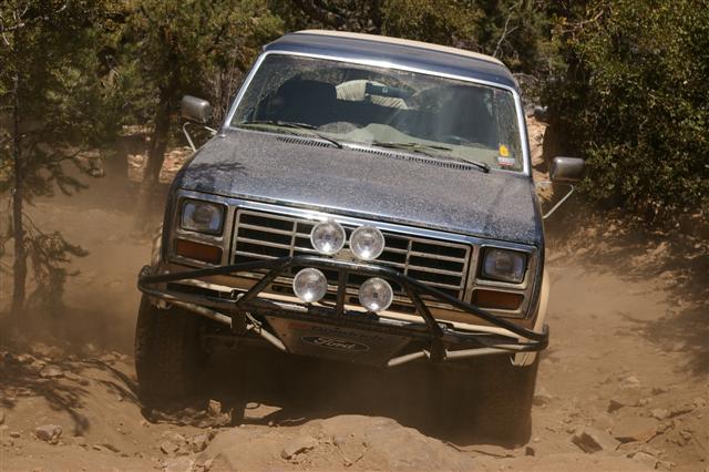 1986 Ford Bronco driving on a dusty, rocky trail with trees in the background