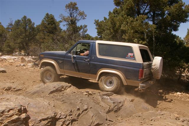 Blue Ford Bronco with tan top driving over rocks on a dirt trail. Spare tire mounted on the back