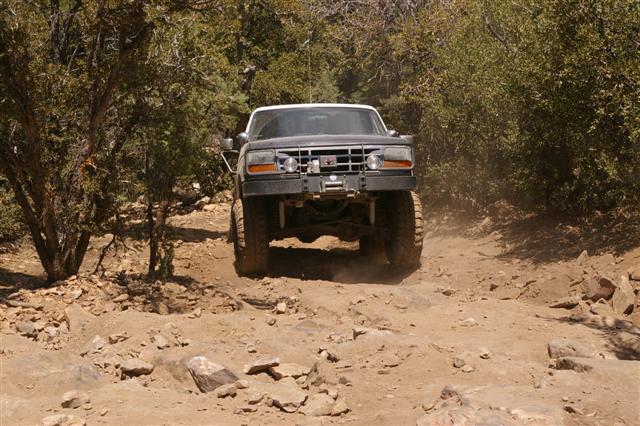 Front view of a lifted 1992 Ford F-150 driving on a rocky dirt trail with trees on either side