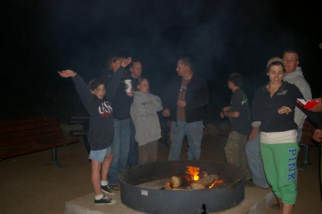 Group of people gathered around a campfire at night, some with arms raised