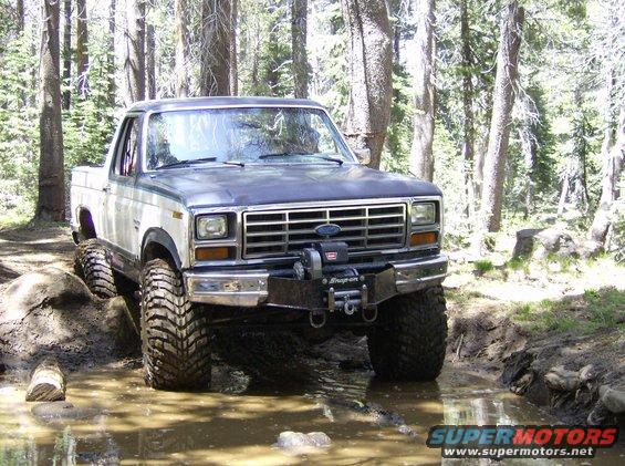 Front view of a modified 1980s Ford pickup truck with large tires and a winch, driving through mud