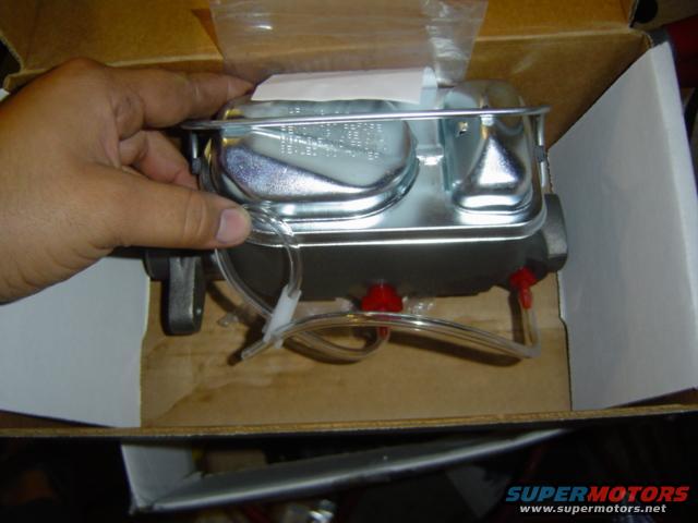 Hand holding a new master cylinder in its box. It has two reservoirs and red caps on the ports