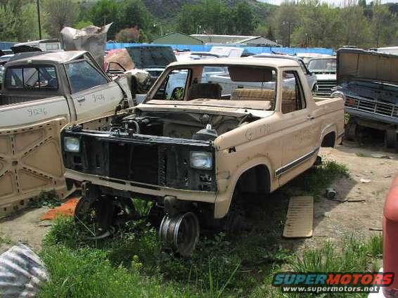 Tan 1980s Ford Bronco with hood open, missing front wheels, in a junkyard. Another truck is to the left