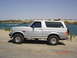 1996 Ford Bronco - This SUV is parked on a sandy area next to a body of water under a clear blue sky.