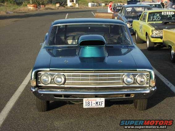 Front view of a blue 1966 Ford Fairlane with a hood scoop and aftermarket wheels. Other cars are visible in the background